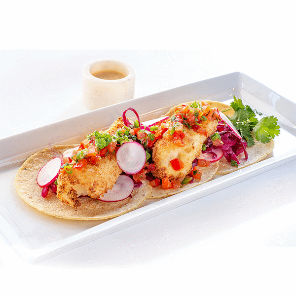 A vibrant culinary presentation featuring three fish tacos on a rectangular white plate, set against a bright white background. Each taco consists of a soft, handmade corn tortilla cradling a golden-brown crispy cod filet, coated with panko breadcrumbs. The cod is topped with a lustrous red cabbage slaw in a deep purple hue, and garnished with thinly sliced radishes, chopped green onions, and fresh cilantro leaves. Accompanying the plate is a small ramekin filled with creamy lemon paprika aioli.